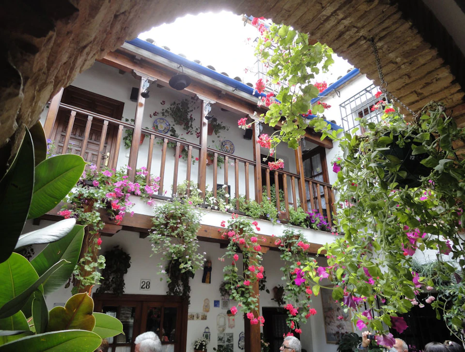 A courtyard with a wooden balcony adorned with vibrant flowers and hanging plants, framed by an archway.