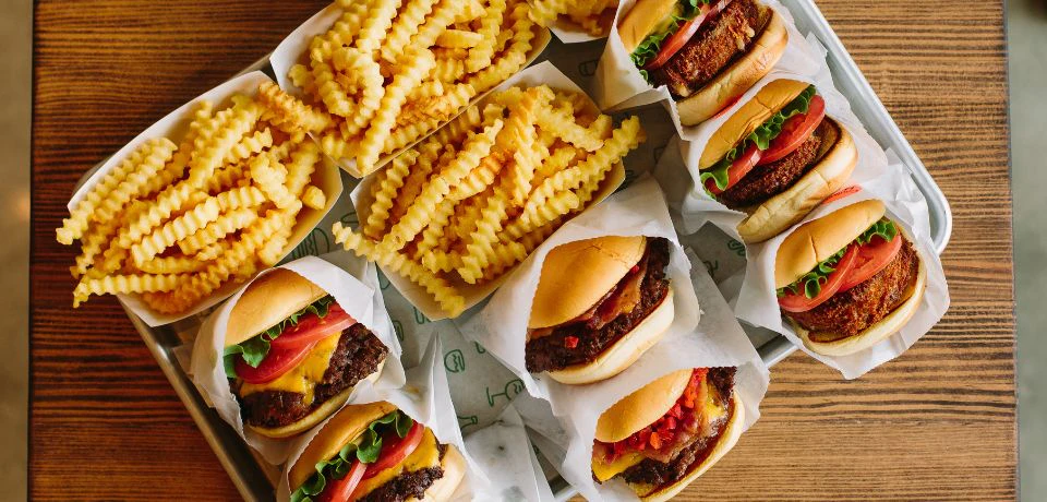 A collection of cheeseburgers with toppings like lettuce and tomato, accompanied by crinkle-cut fries on a table.
