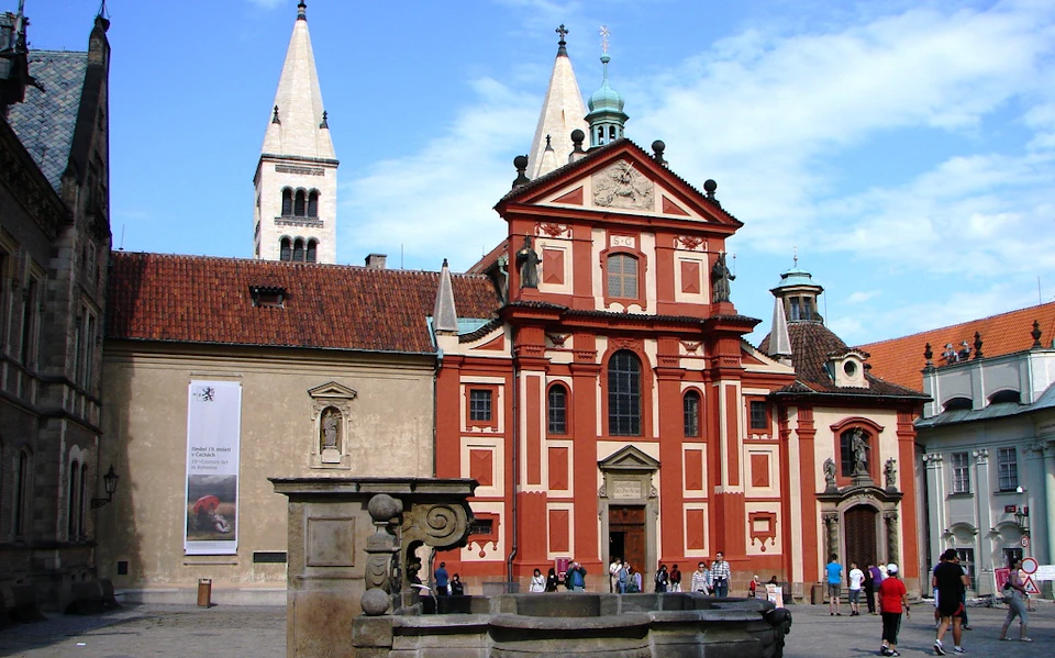 Historic church with red and white facade, tall tower, and a fountain in the foreground, with people walking in front.