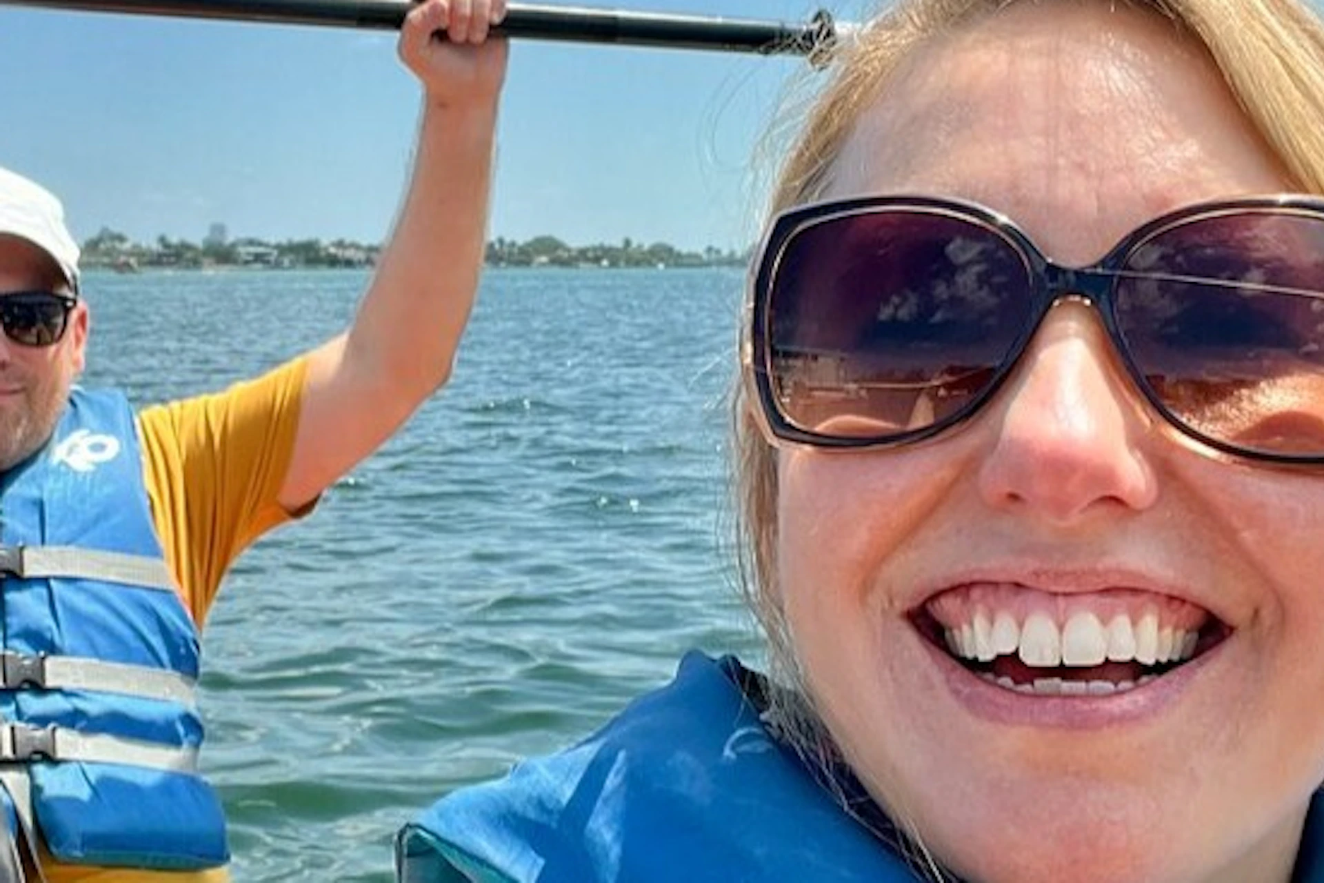 Two people kayaking on a sunny day, wearing blue life vests. The person in front smiles at the camera, with water and sky visible.