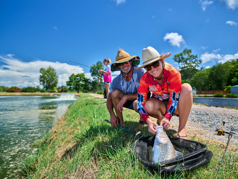 Two people in hats kneel by a pond, smiling and holding a fish in a net. In the background, another person is fishing.