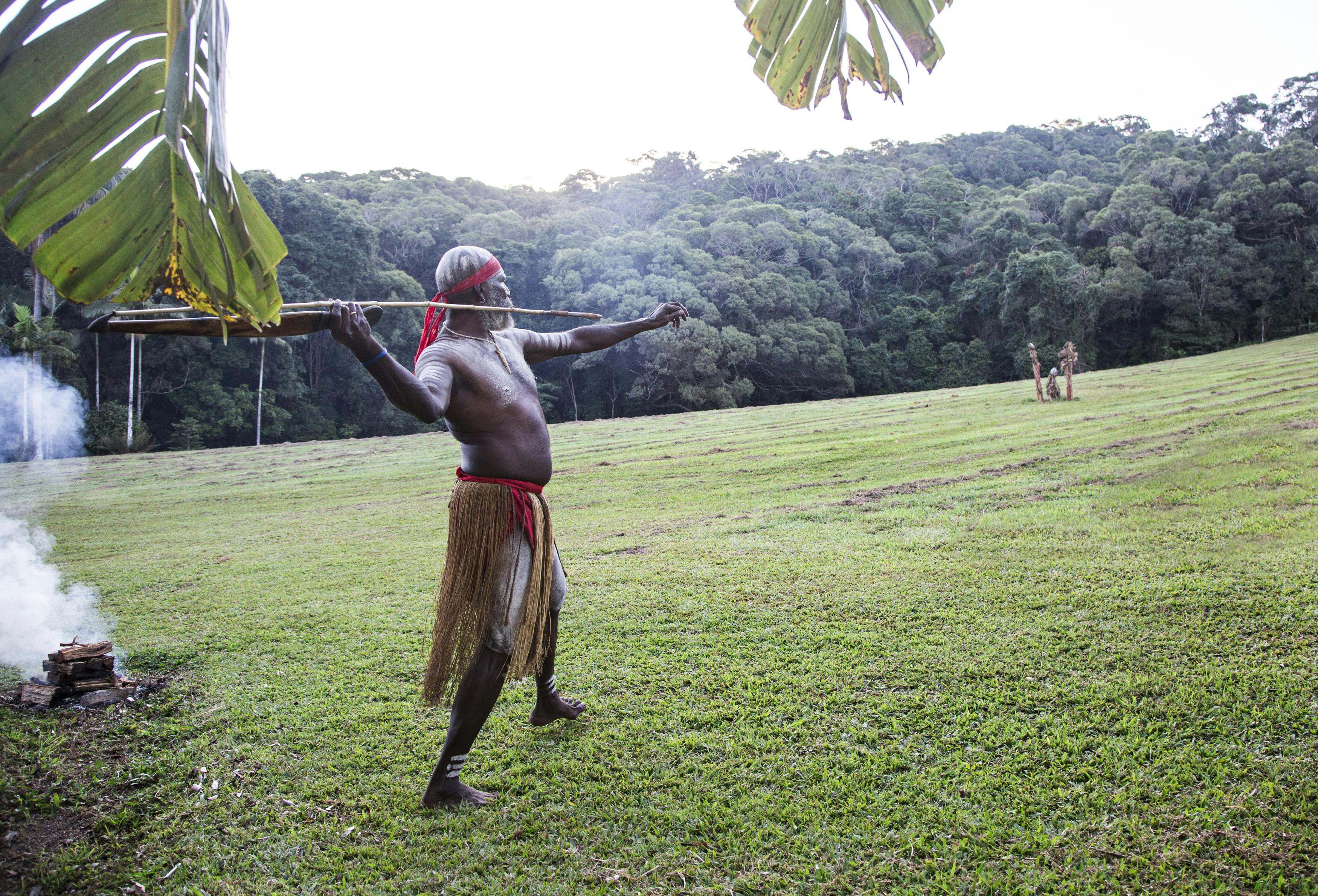 一名身着传统服装的男子站在草地上，拿着一支长矛，瞄准远处的森林。