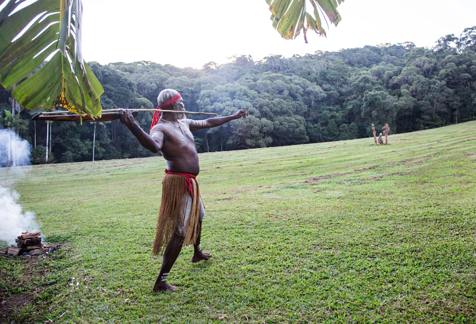 A man dressed in traditional clothing stands on a grassy field, holding a spear aimed at a distance with forested hills in the background.