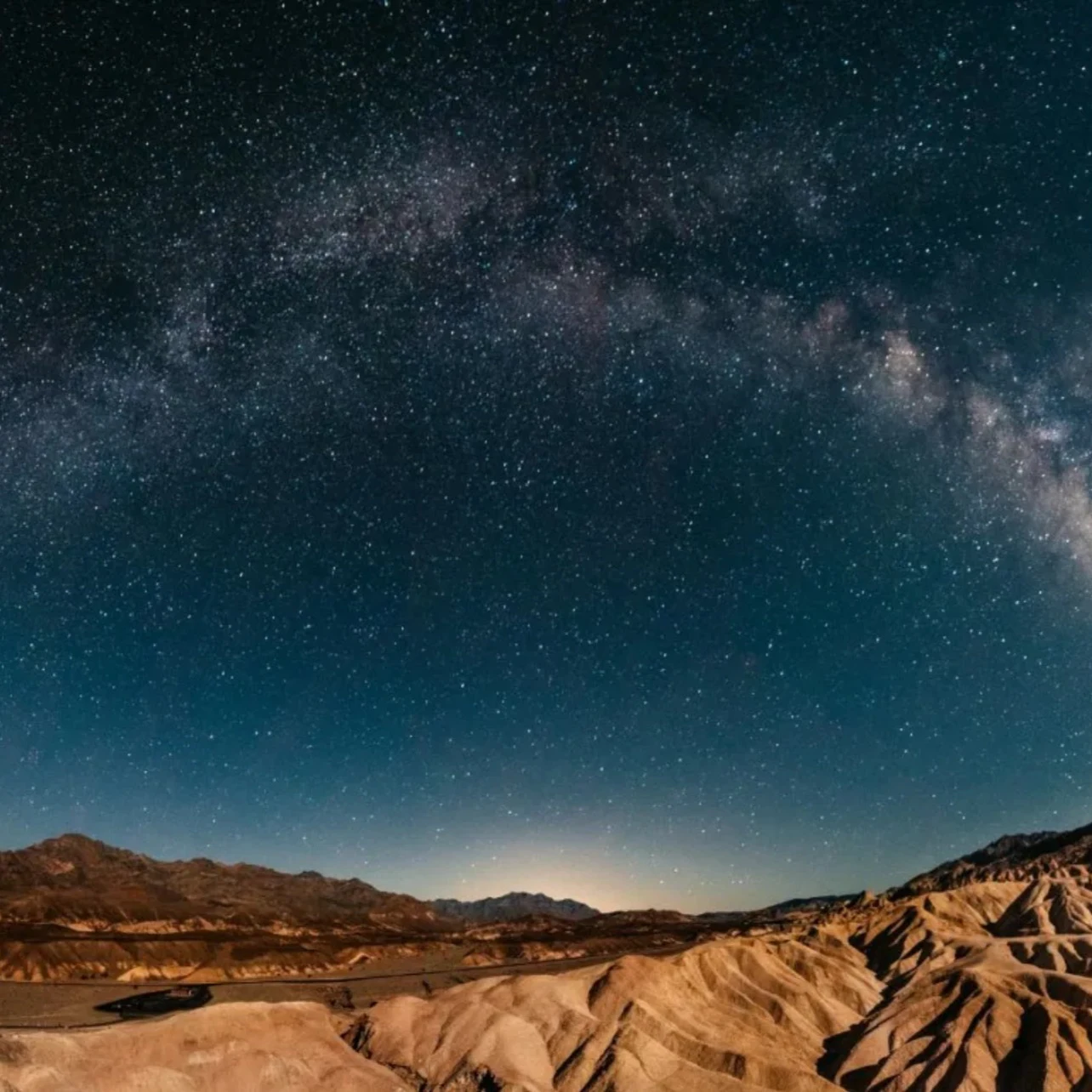 Panoramic night view of a desert landscape with the Milky Way arching across the dark, star-filled sky.