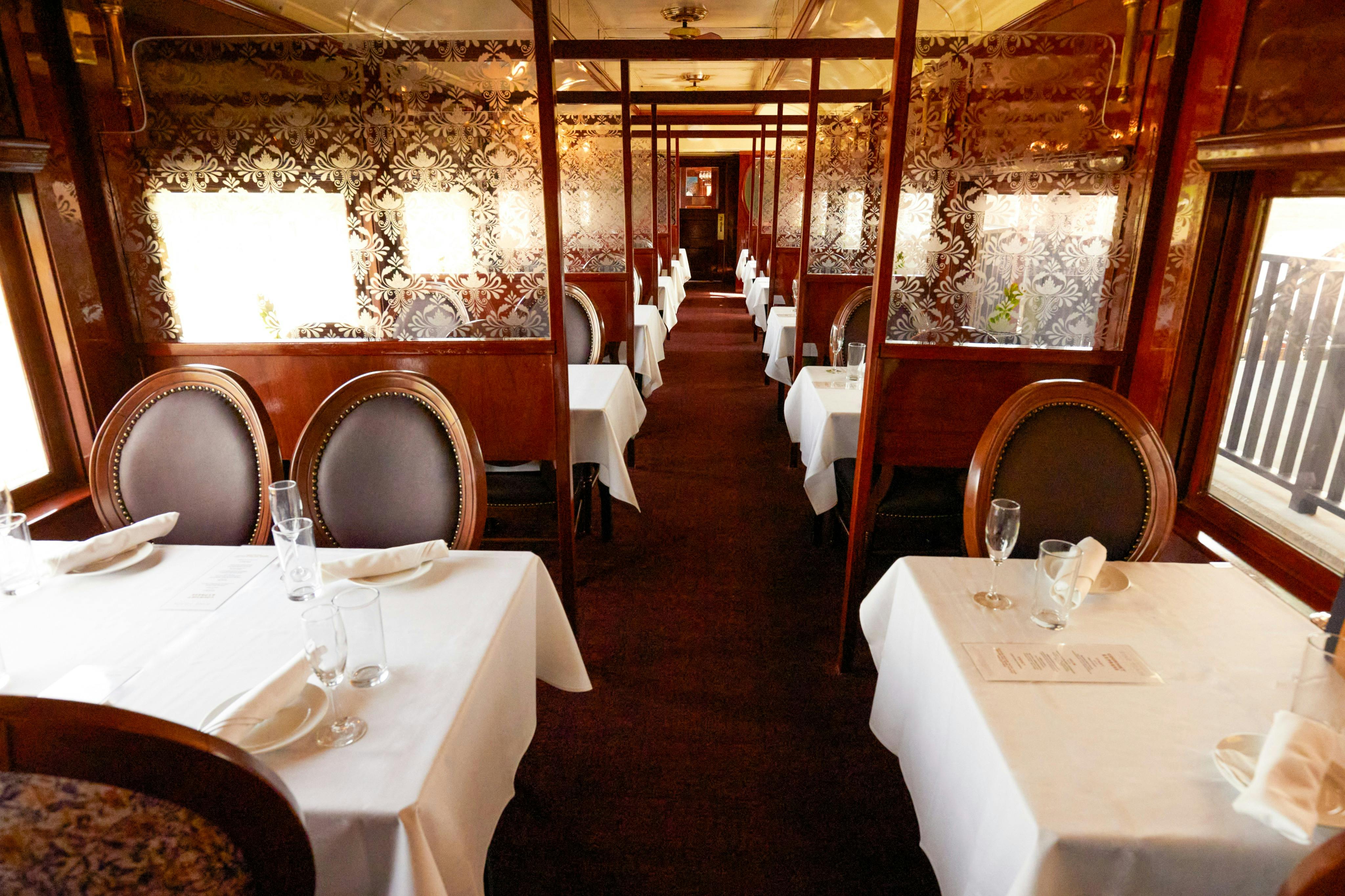 An elegant train dining car with tables set with white linens and chairs, featuring ornate floral-patterned walls.