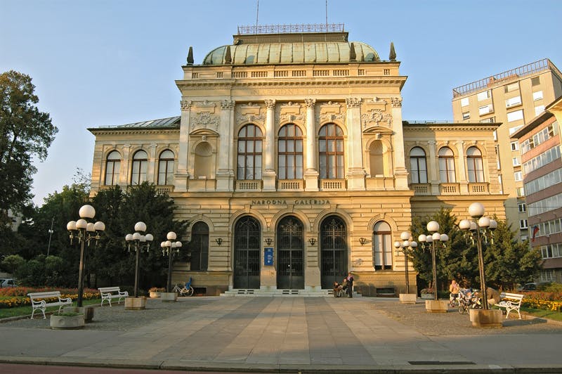 Historická budova s klenutými okny a zelenou střechou, označená jako „Narodna Galerija“, přiléhající k moderní budově.