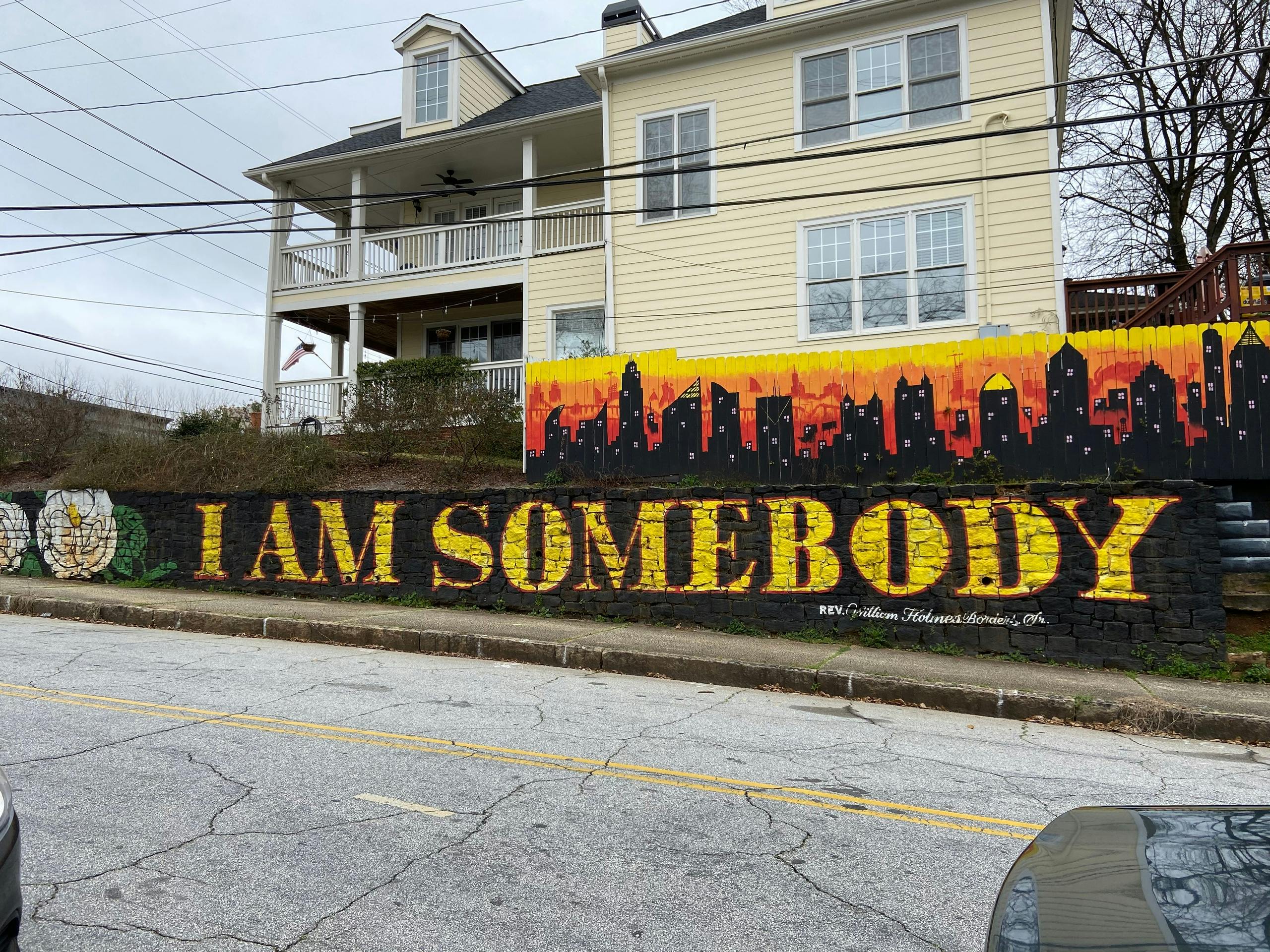 Une peinture murale avec le texte "I AM SOMEBODY" en grandes lettres jaunes sur un mur, avec une ligne d'horizon de la ville et une maison en arrière-plan.