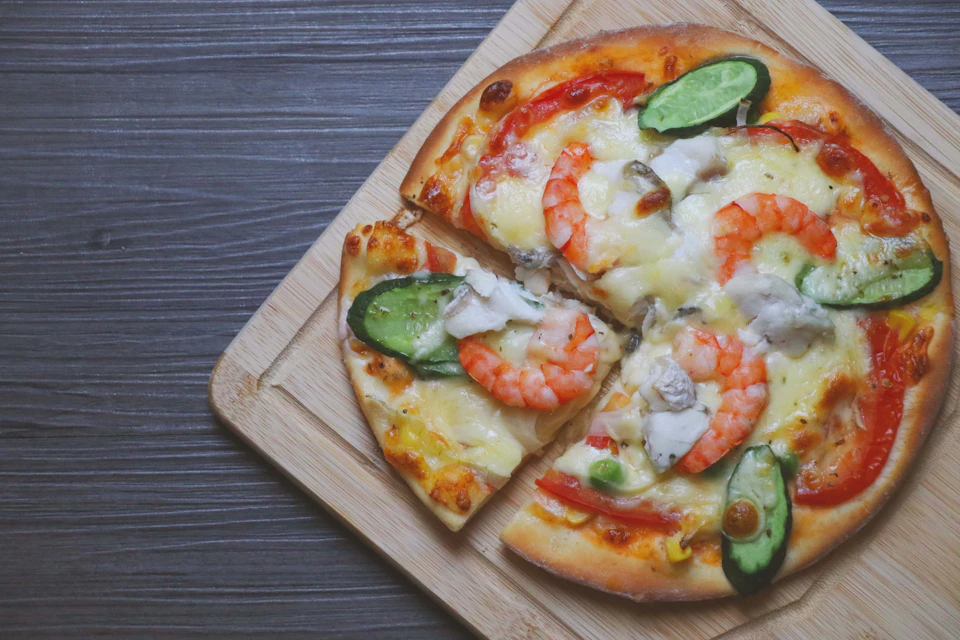 A seafood pizza with shrimp, vegetables, and cheese on a wooden serving board, placed on a gray wooden surface.