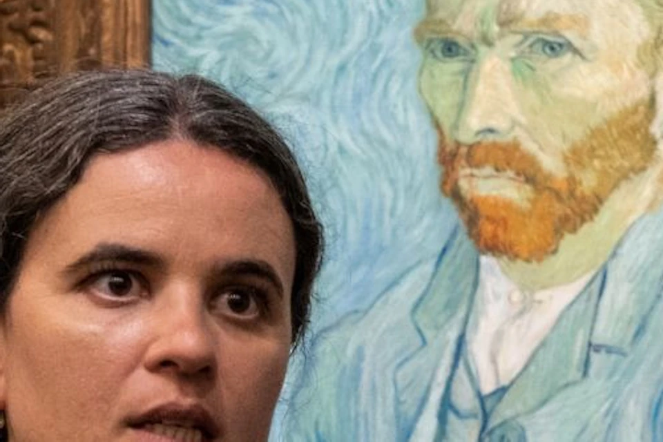 A person gesturing while speaking near a framed painting of a man with a red beard and blue background in a museum setting.