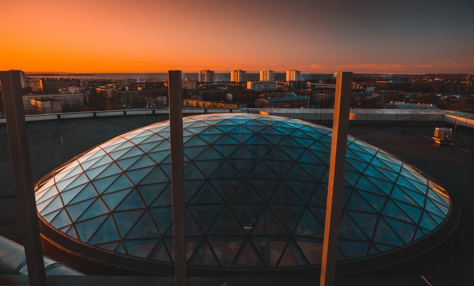 Geodesic glass dome structure on a rooftop at sunset, with city buildings and a colorful sky in the background.