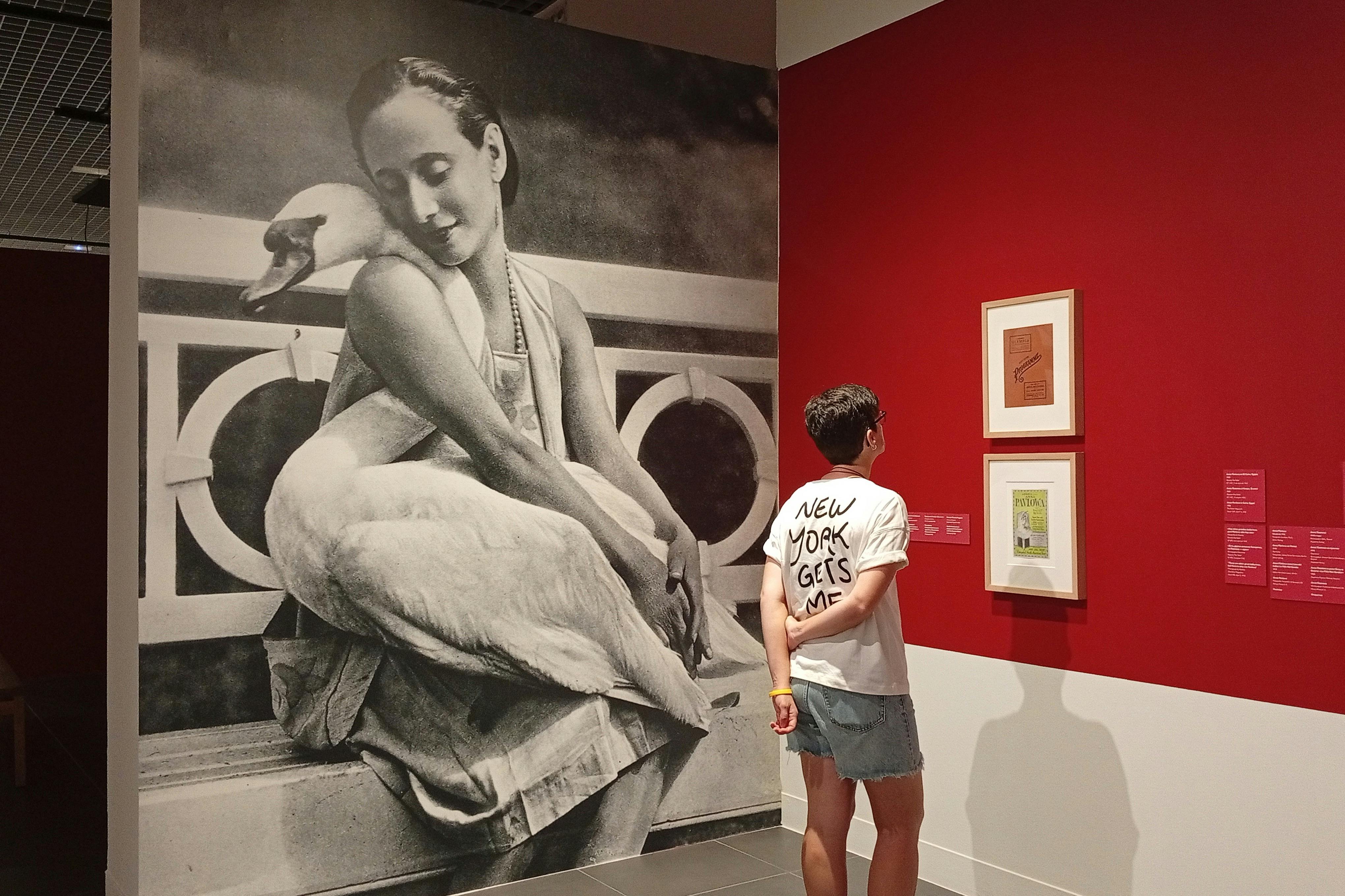 Um grande mural a preto e branco de uma mulher a segurar um cisne está exposto numa parede. Uma pessoa de camisa branca está por perto.