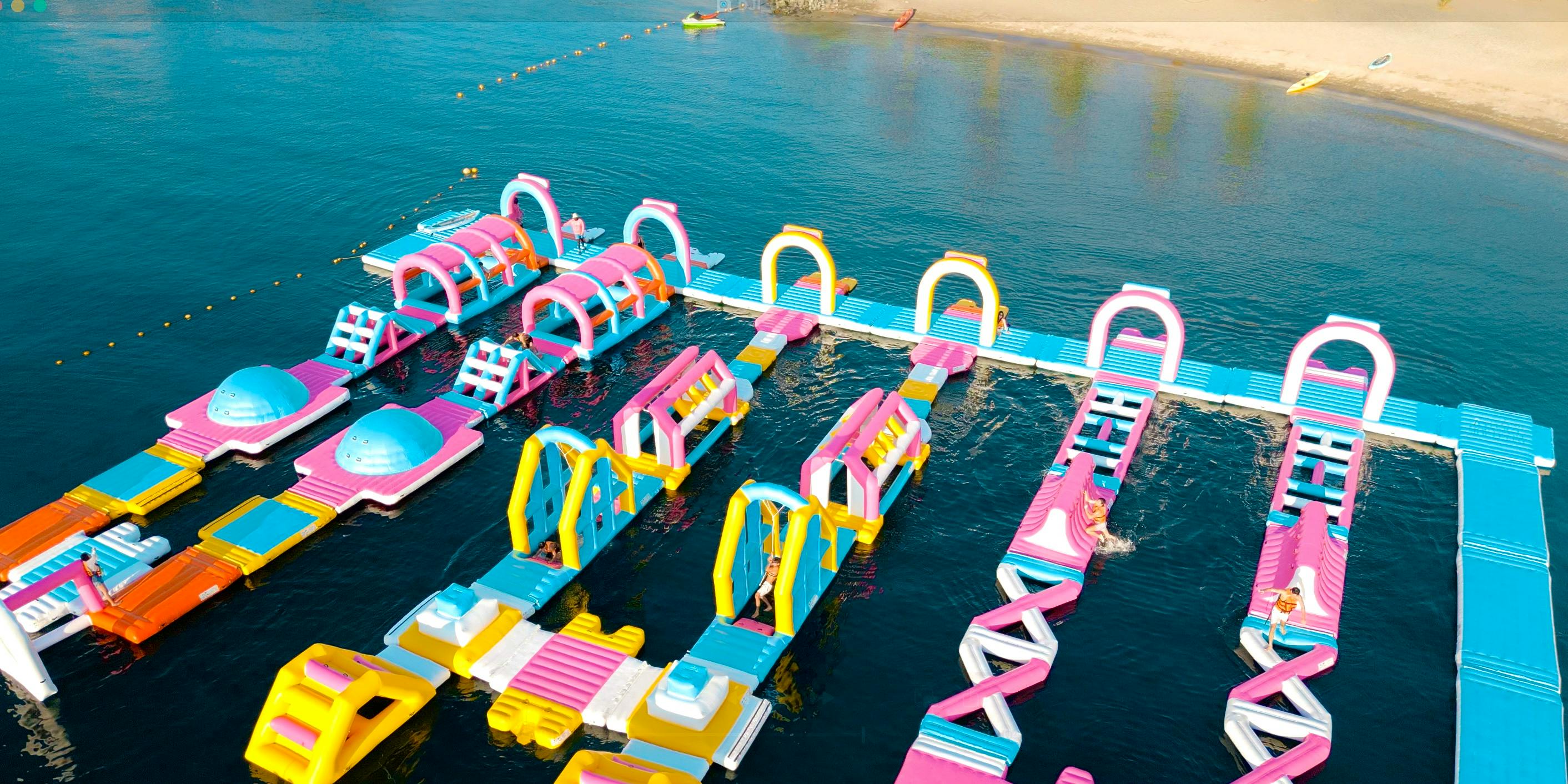 A colorful inflatable water park with various obstacles and slides, floating on a calm blue body of water near a sandy beach.