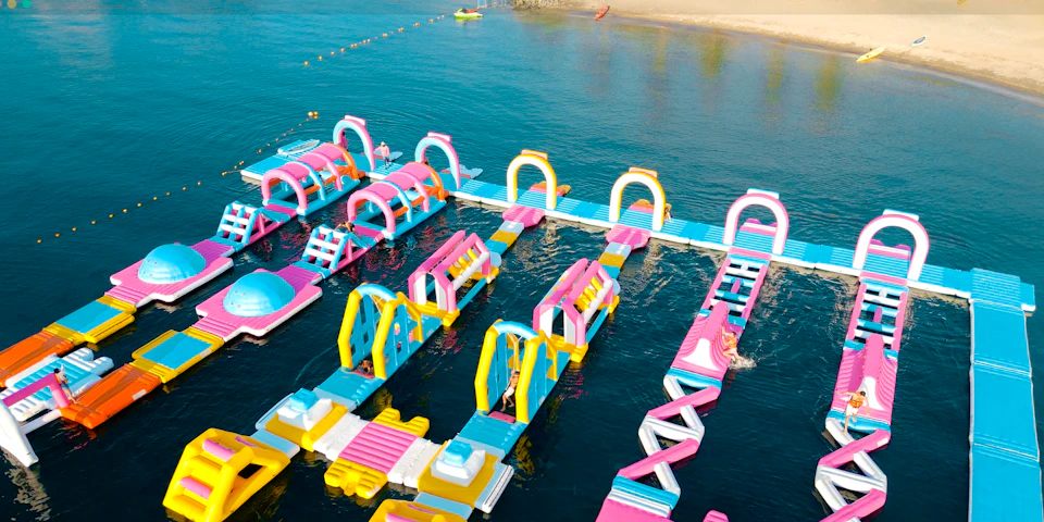A colorful floating inflatable water park on a calm body of water near a sandy beach, with various slides and obstacles.