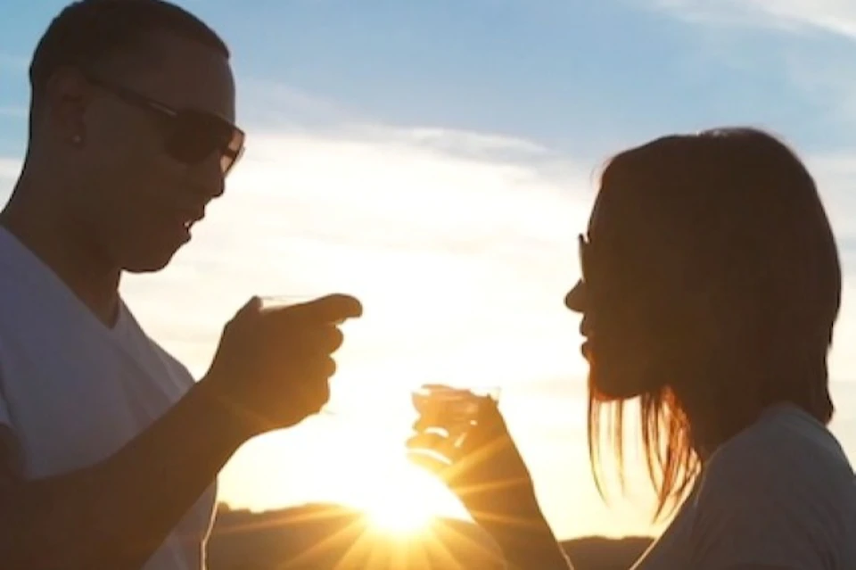 Silhouettes of two people wearing sunglasses, holding drinks and gesturing against a sunset background.