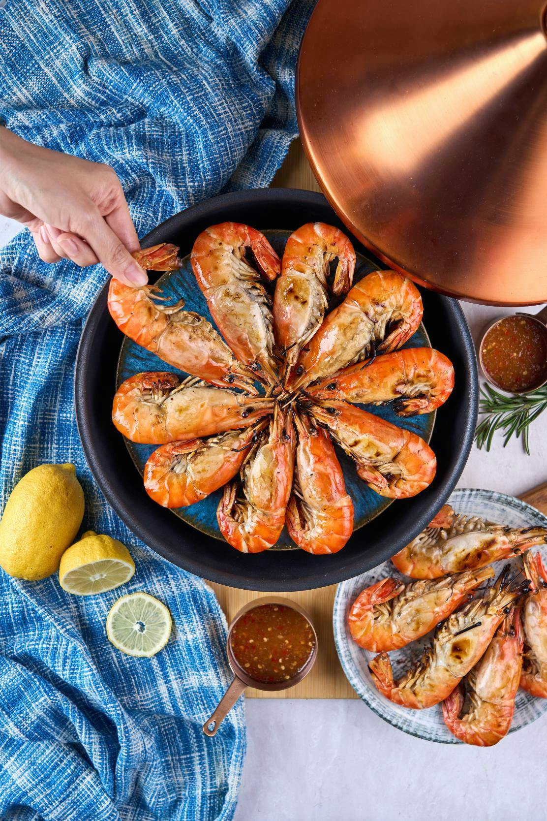 A plate of grilled prawns arranged in a circular pattern, with lemons, sauces, and a hand lifting the cloche nearby.