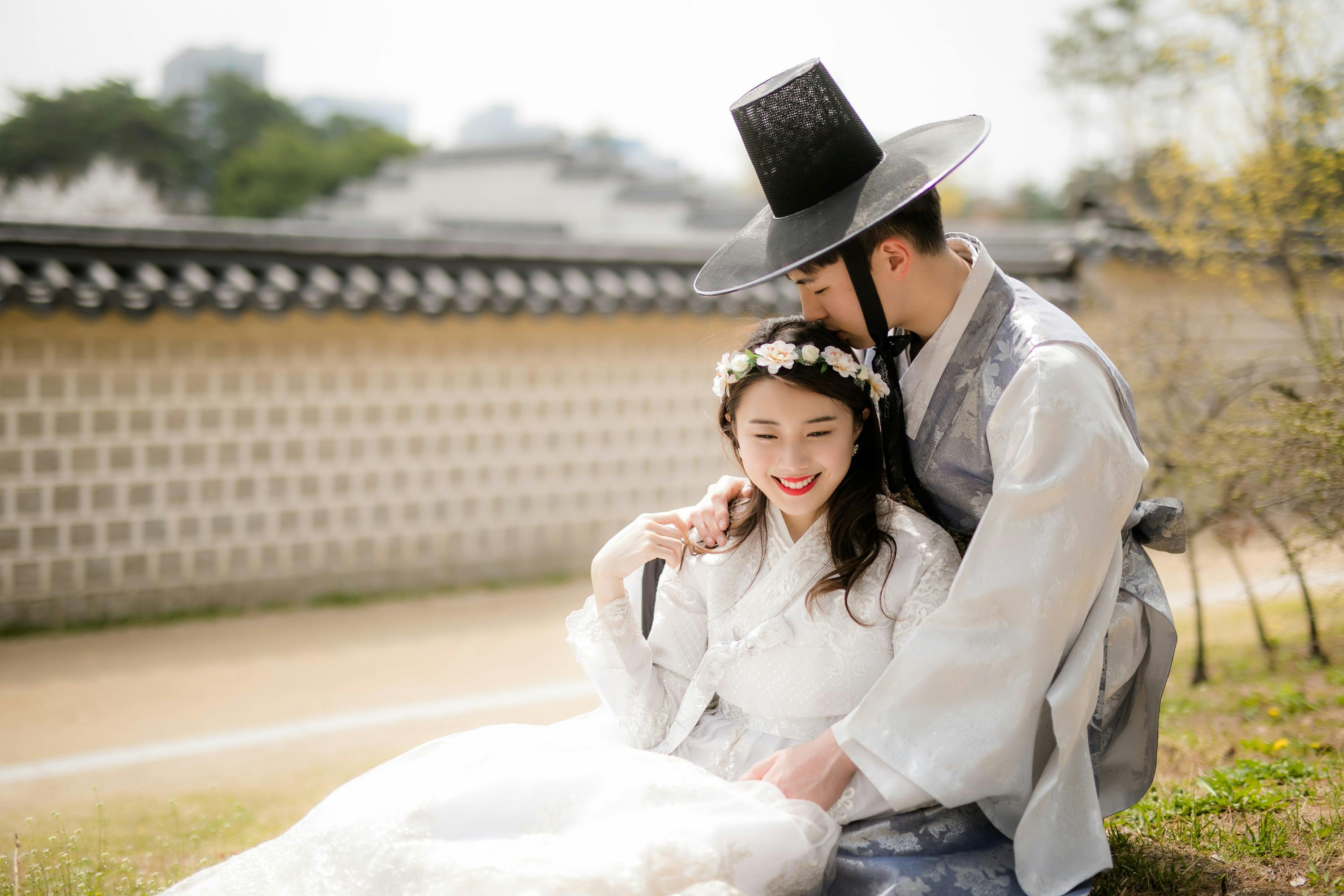 A couple dressed in traditional Korean hanbok, with the man embracing the woman against a background of a wall and trees.