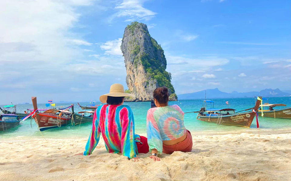 Two people in colorful shirts sit on a sandy beach facing the ocean, with boats and a tall rock formation in the background.