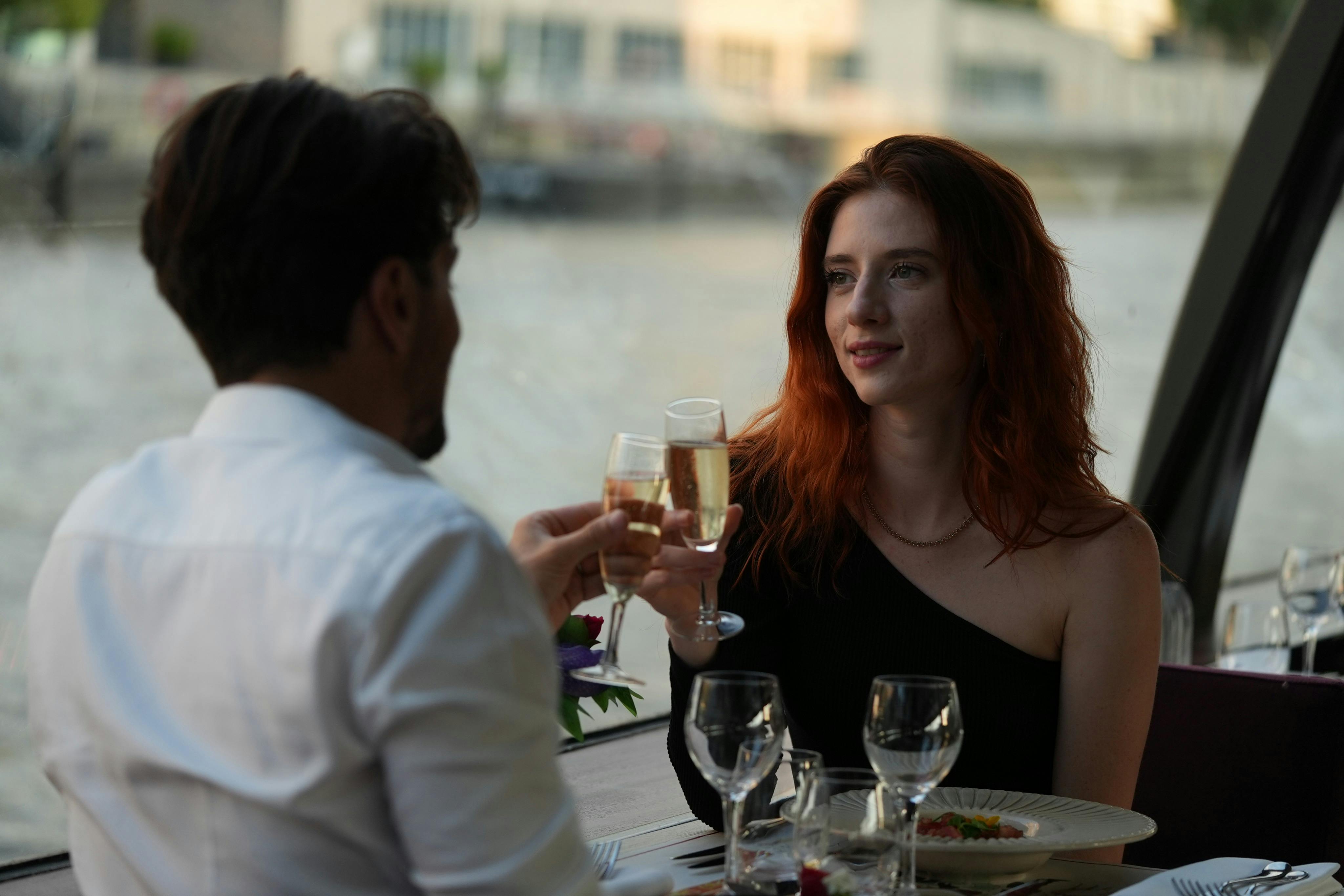 Eine Frau mit rotem Haar und schwarzem Oberteil sitzt an einem Esstisch mit Weingläsern und stößt mit einer anderen Person an.