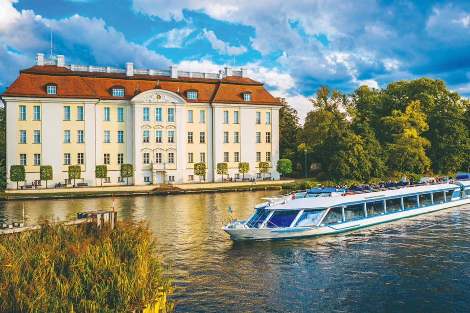 A boat travels on a river past a large, historic building with a red roof, bordered by trees and greenery under a partly cloudy sky.