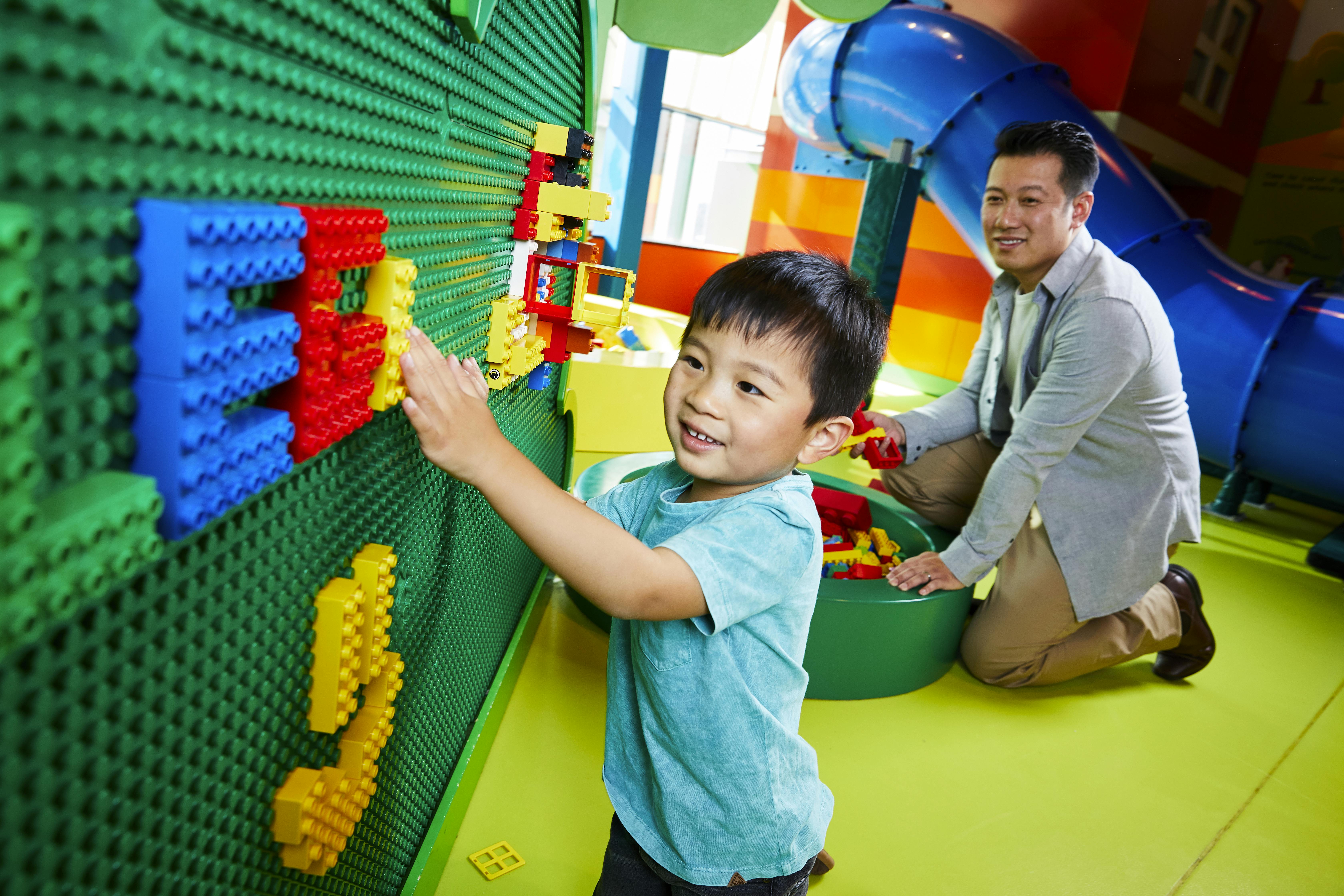 A smiling child plays with building bricks on a green wall while a man kneels nearby, assembling bricks. Blue slide in background.