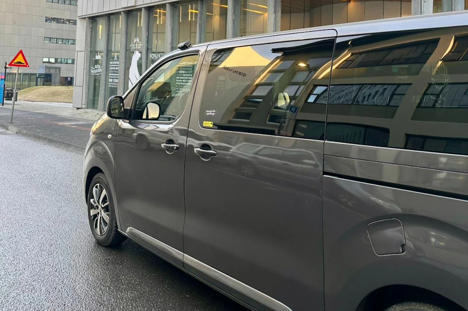 A gray minivan is parked on a wet street in front of a modern building with large windows and visible reflections.