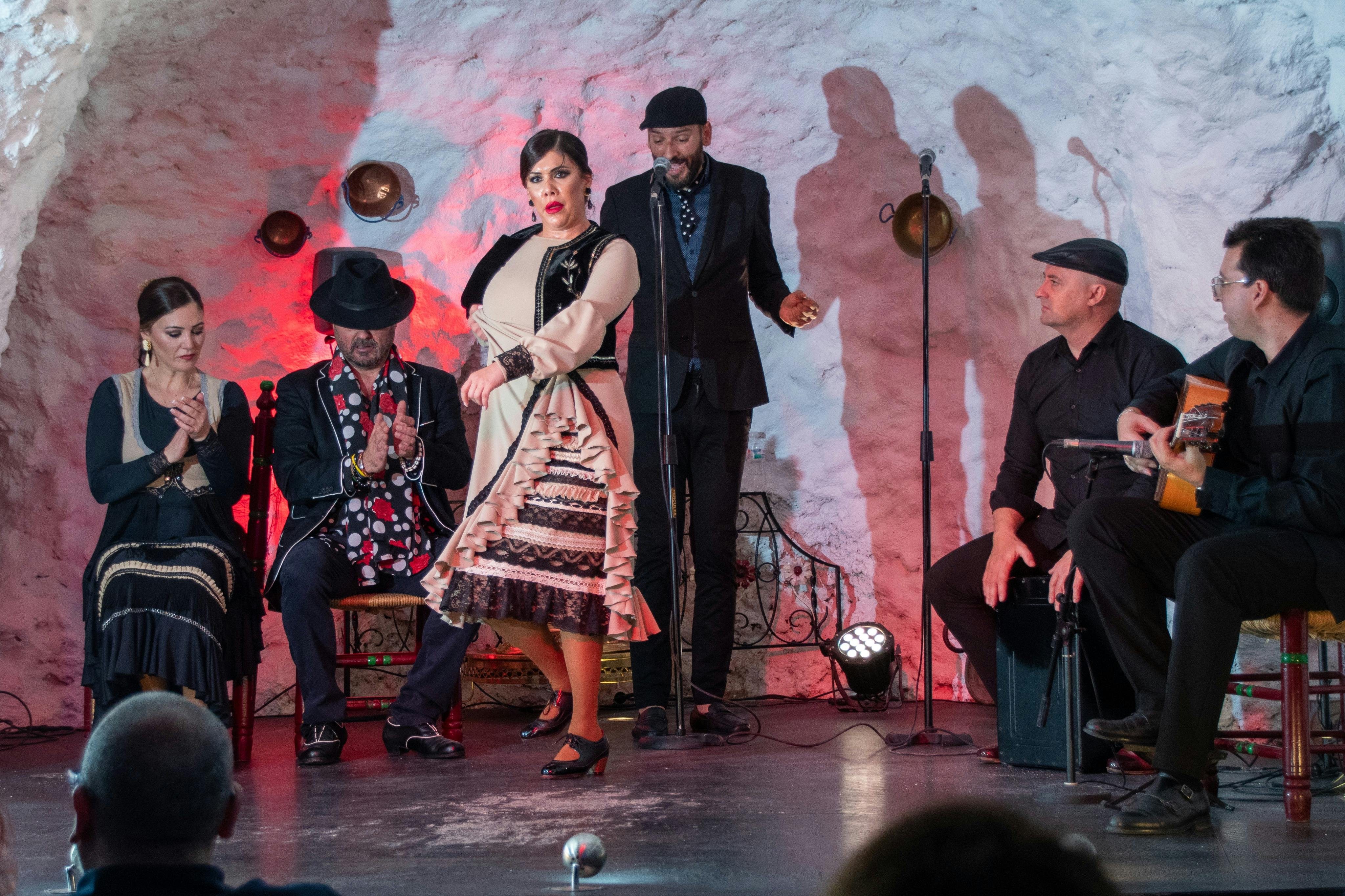 Four flamenco performers, including two seated and two standing, perform in a cave-like setting with red lighting accents.