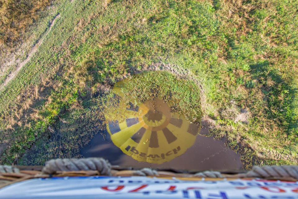 View from a hot air balloon showing its shadow on a grassy and marshy landscape below.