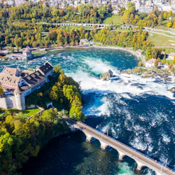 Flygfoto över en stor flod med ett slott, pulserande höstträd och en bro över tumultartat vatten som leder till en liten stad.