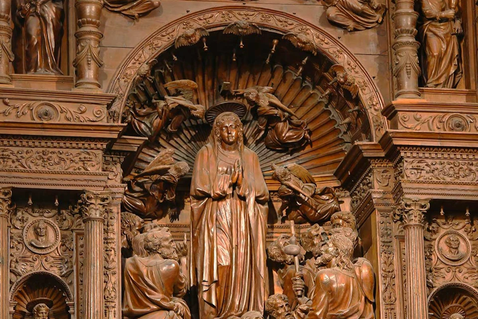 Intricate wooden sculpture depicting a central figure surrounded by angels and people, with detailed carvings and arches.