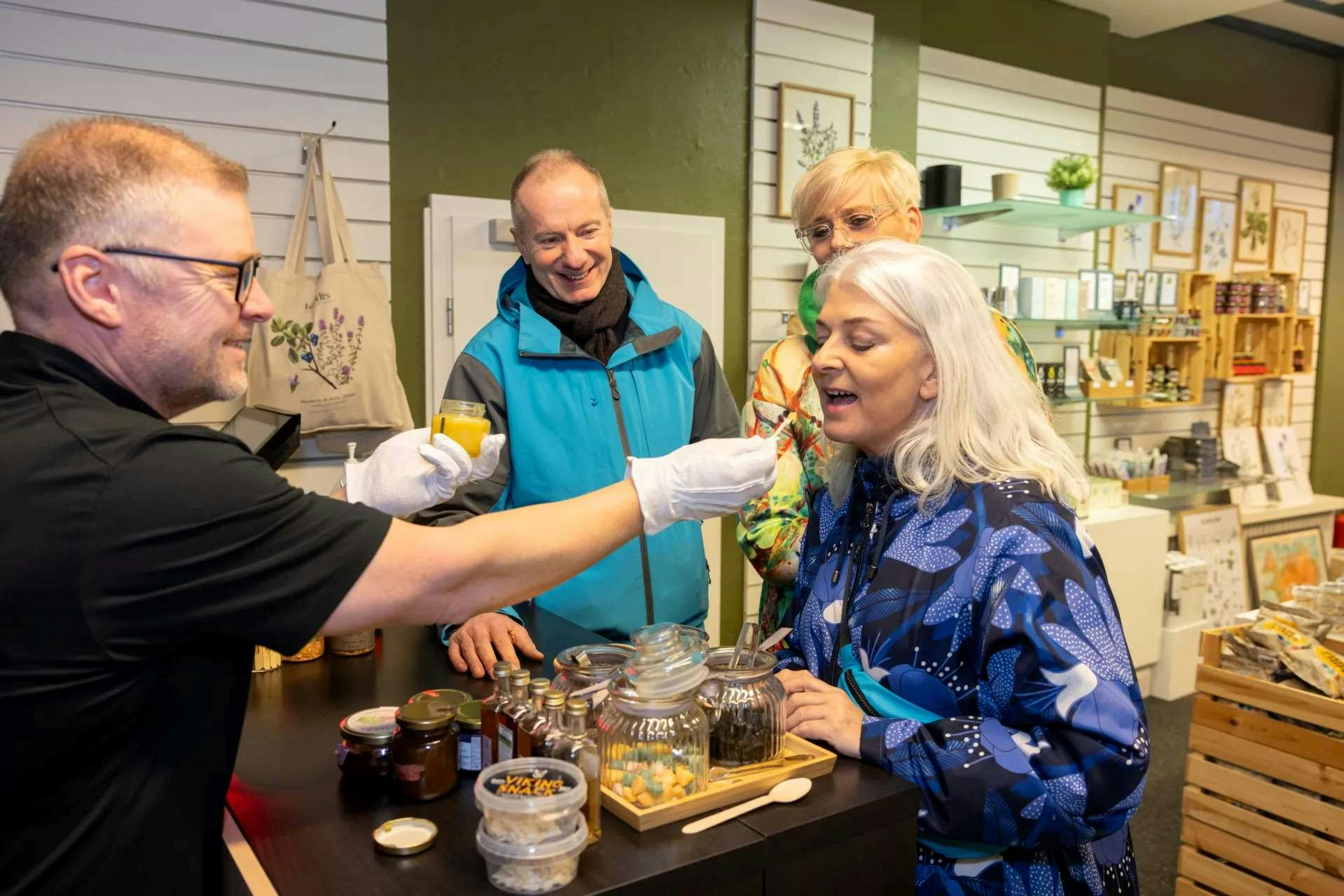Three people taste samples offered by a person in gloves at a shop with jars and framed art on shelves.