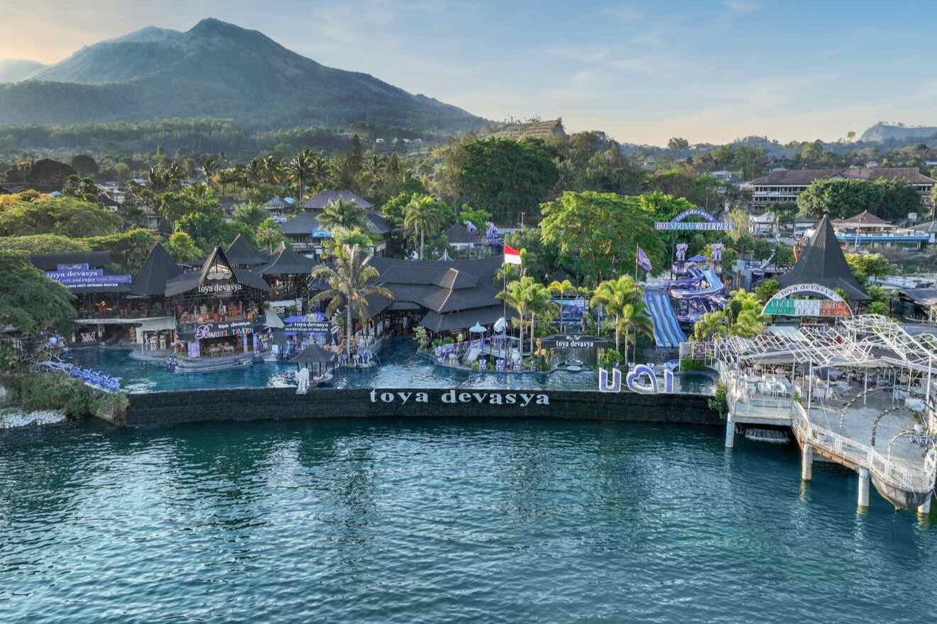 An aerial view of Toya Devasya resort with a pool, palm trees, and buildings, set against a mountainous backdrop.
