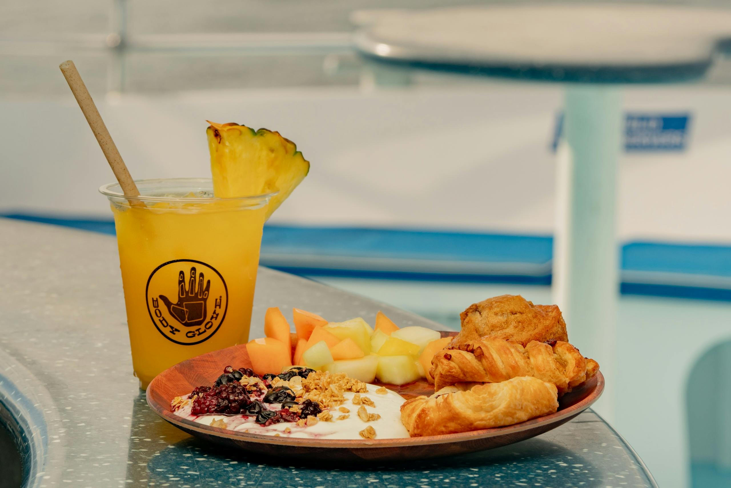 A plate with yogurt, granola, fruit, and pastries, alongside a pineapple-topped yellow drink, on a table.