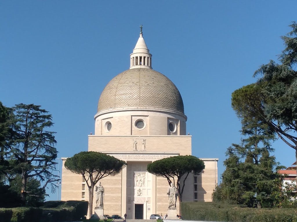 Uma grande igreja em cúpula com estátuas, ladeada por árvores, num céu azul e límpido.