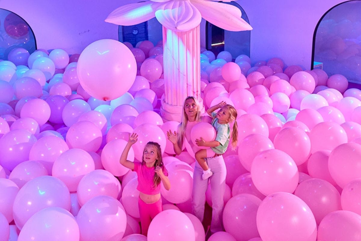 한 여성이 분홍색 풍선으로 둘러싸인 아이를 안고 있으며, 옆에 다른 아이가 커다란 기둥이 있는 분홍색 조명 방에 있습니다.