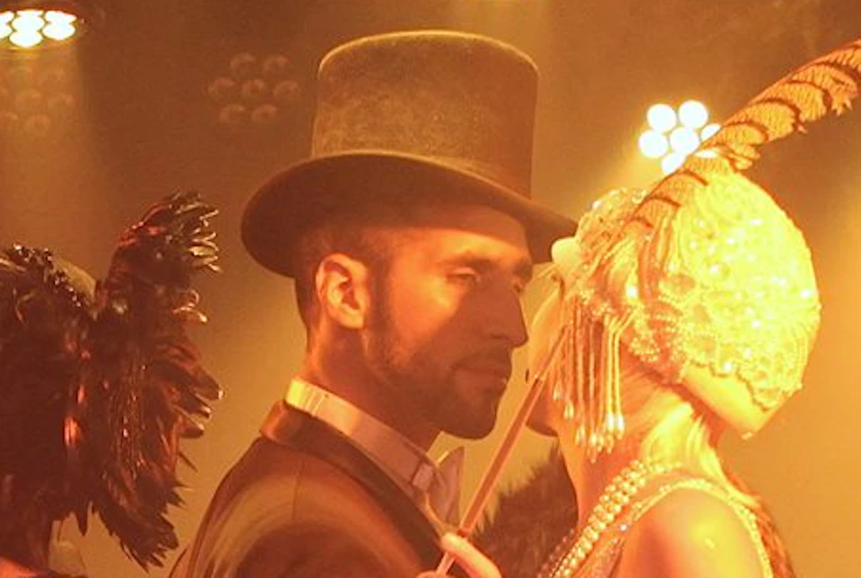 People dressed in 1920s-style costumes, including flapper dresses and top hats, in a dimly lit, elegant room with vintage decor.