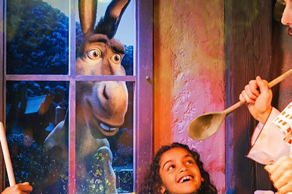 Two girls and a man stir a cauldron emitting green light, while a cartoon donkey looks through the window behind them.
