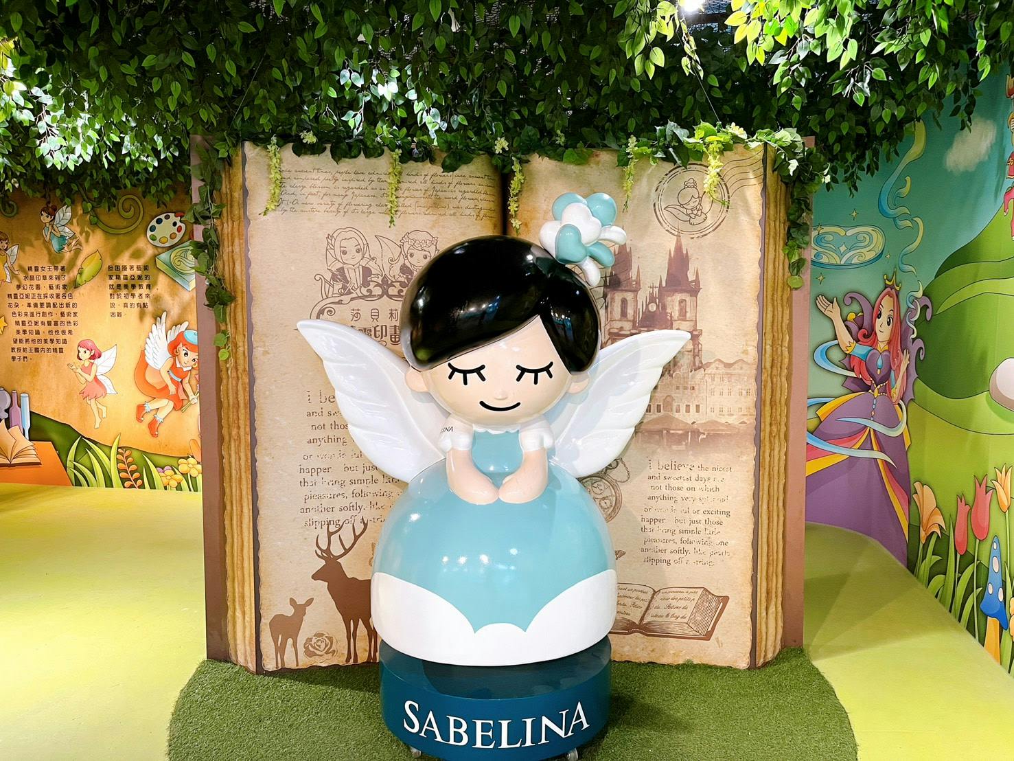 A statue of a cartoon angel with white wings and a blue dress, standing in front of an open book with illustrations.