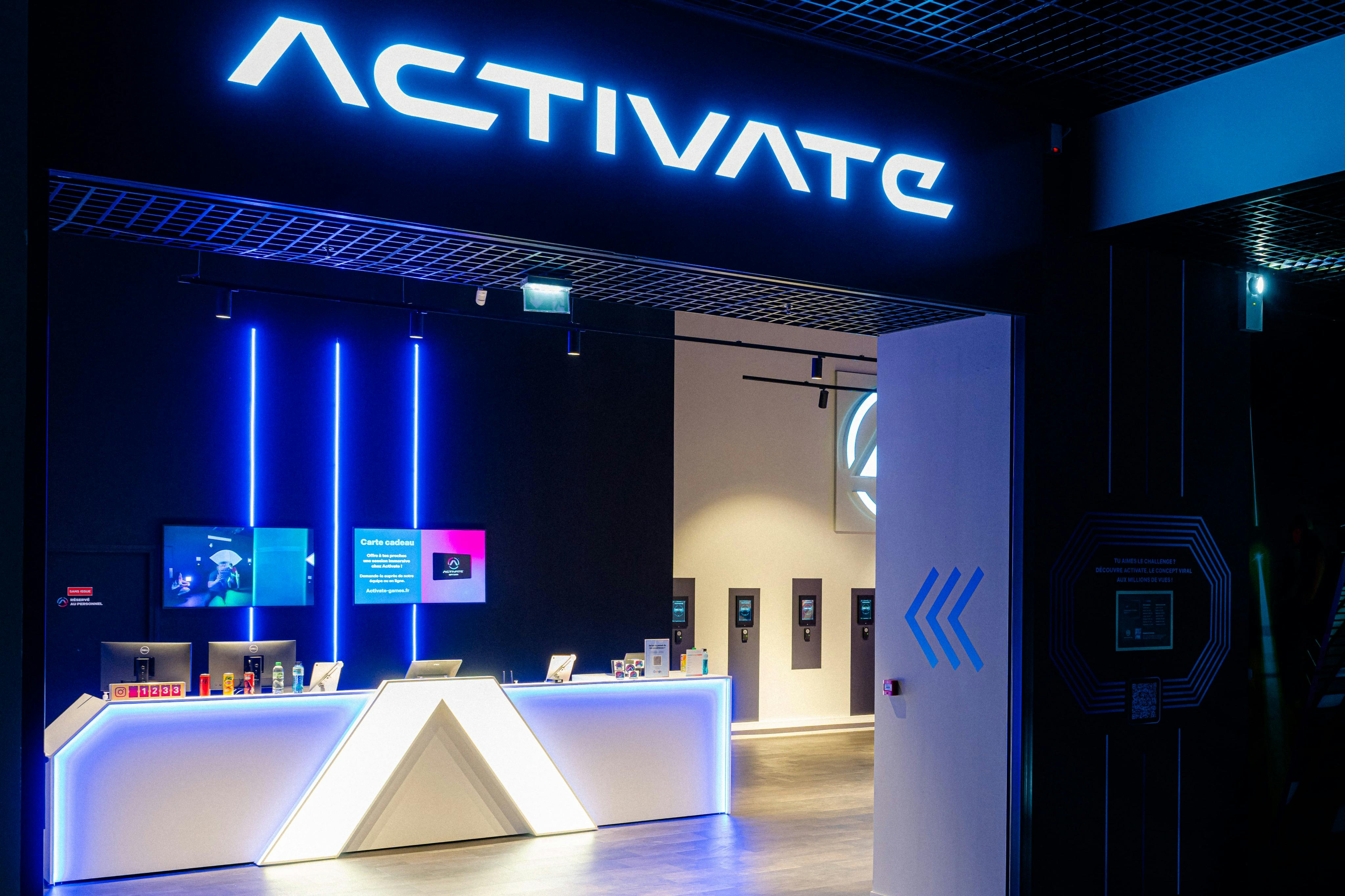빛나는 "Activate" 표지판, 조명 카운터, 벽에 디지털 스크린이 있는 현대적인 리셉션 공간입니다.