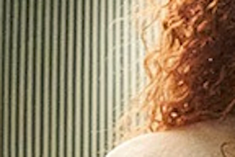 A person with curly hair sits on a stool at a mirrored vanity in a pastel-colored room with a chandelier overhead.
