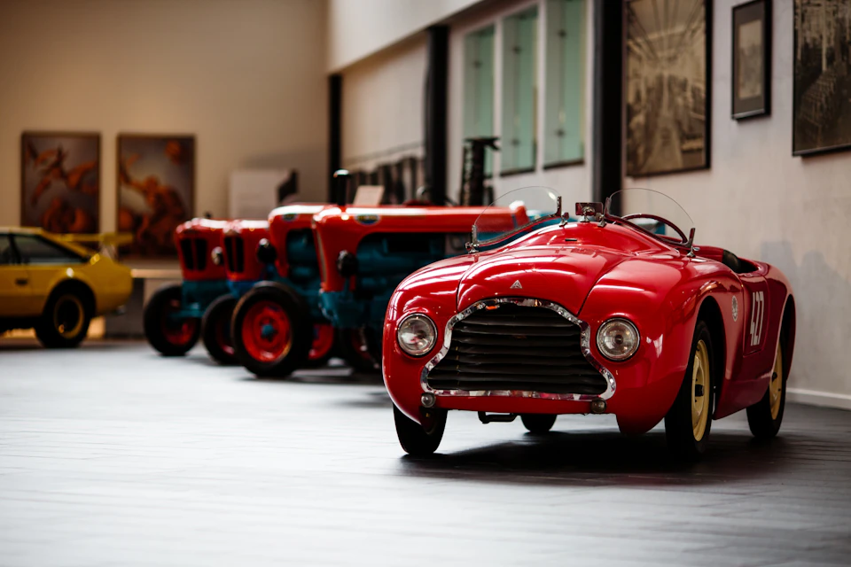 Classic red car displayed in a museum, with several vintage blue and red tractors lined up in the background.
