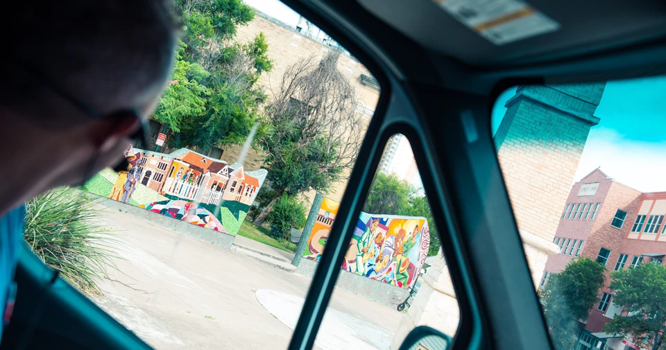 View through a car window of colorful murals and trees in an urban area on a sunny day.