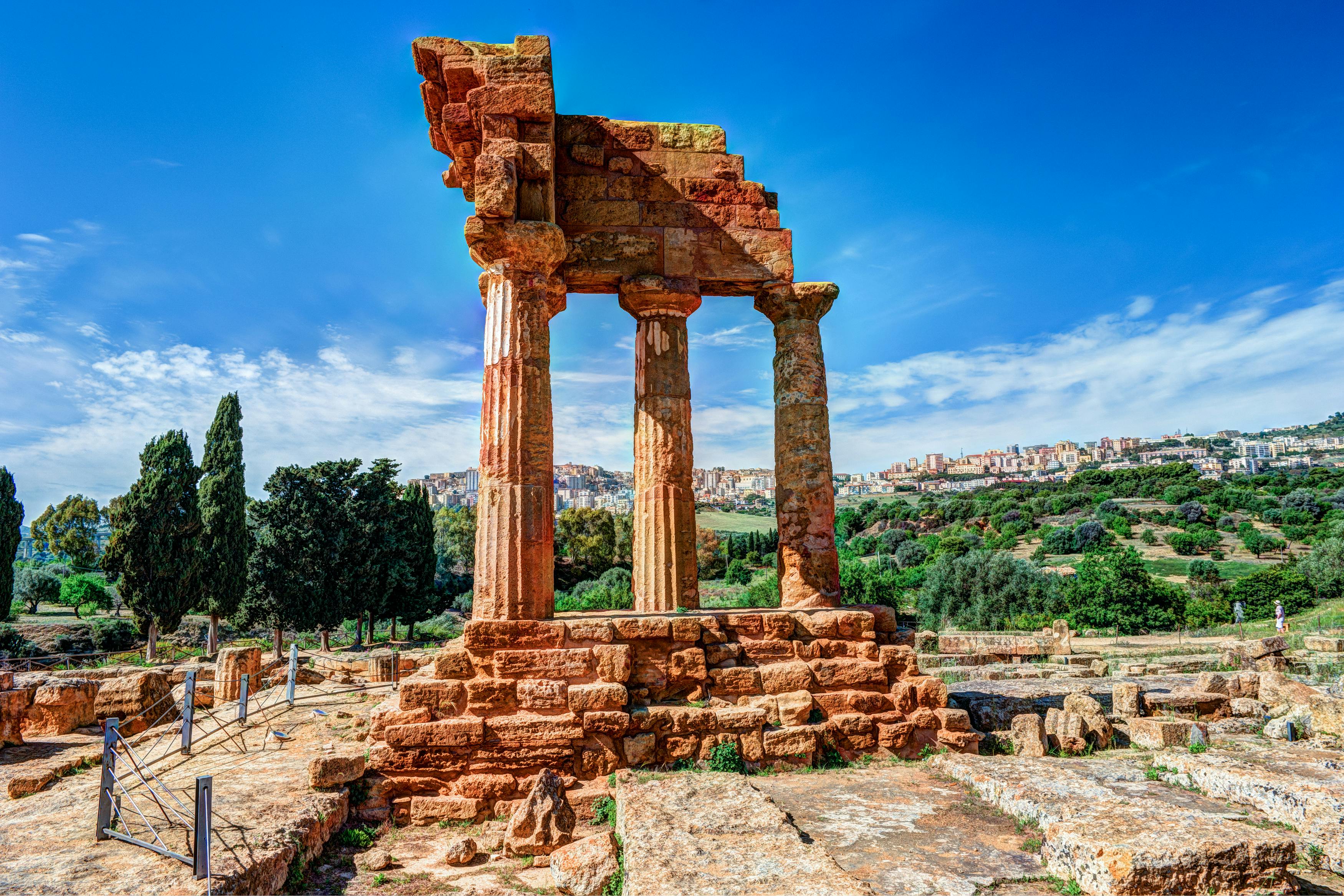 Ancient stone columns and partial structures among ruins, with trees and distant buildings under a bright blue sky.
