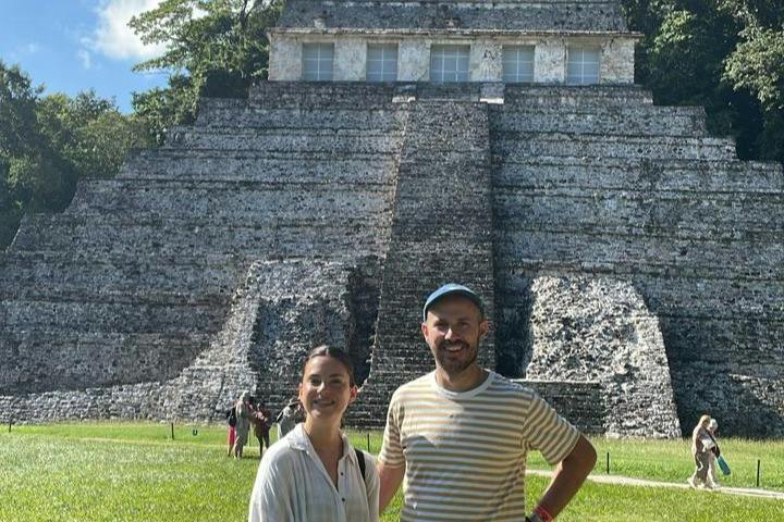 Two people standing in front of an ancient stepped pyramid structure under a sunny sky.