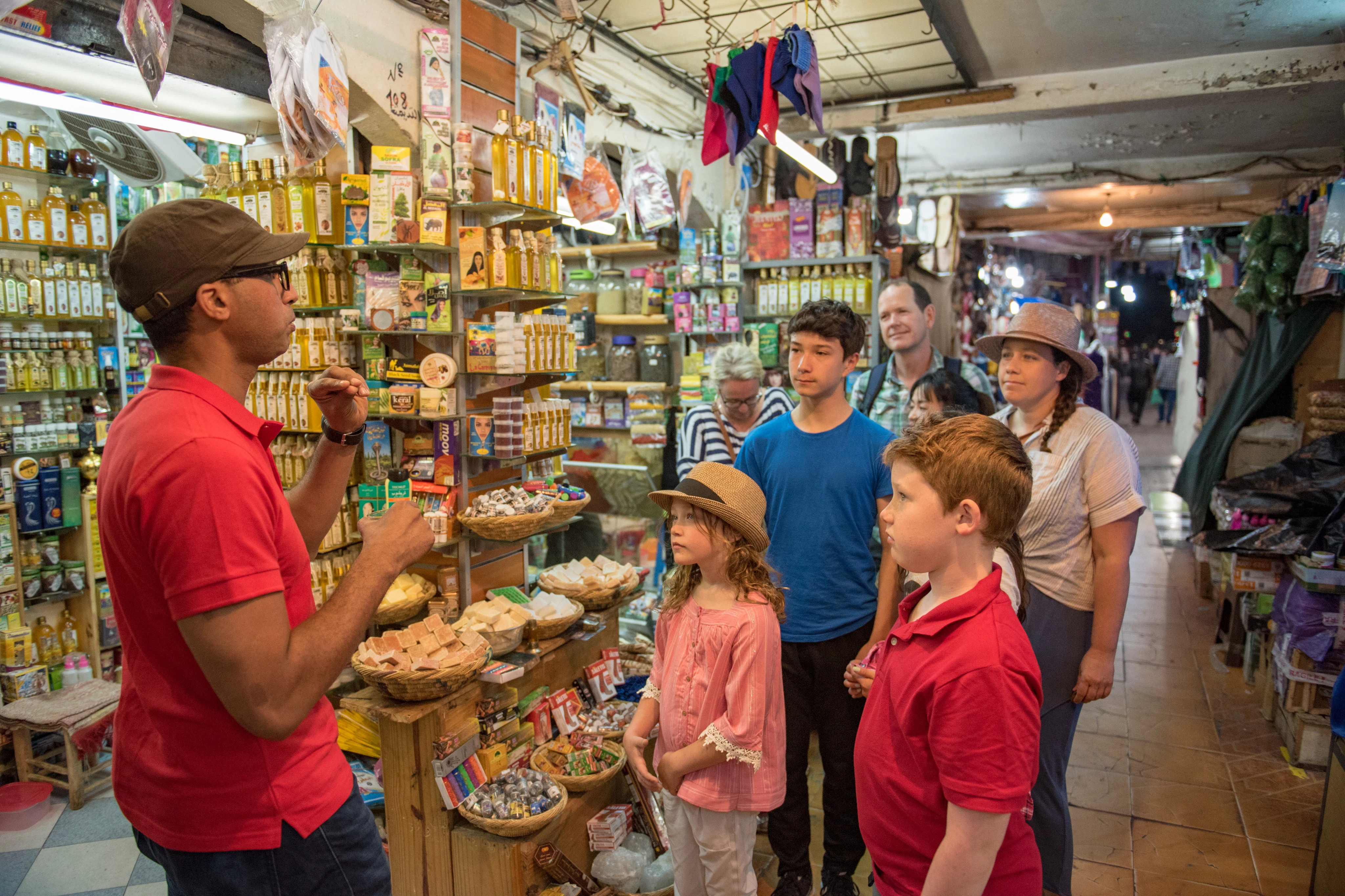 A group of people, including children, stand attentively in a colorful, crowded market shop as a vendor talks to them.