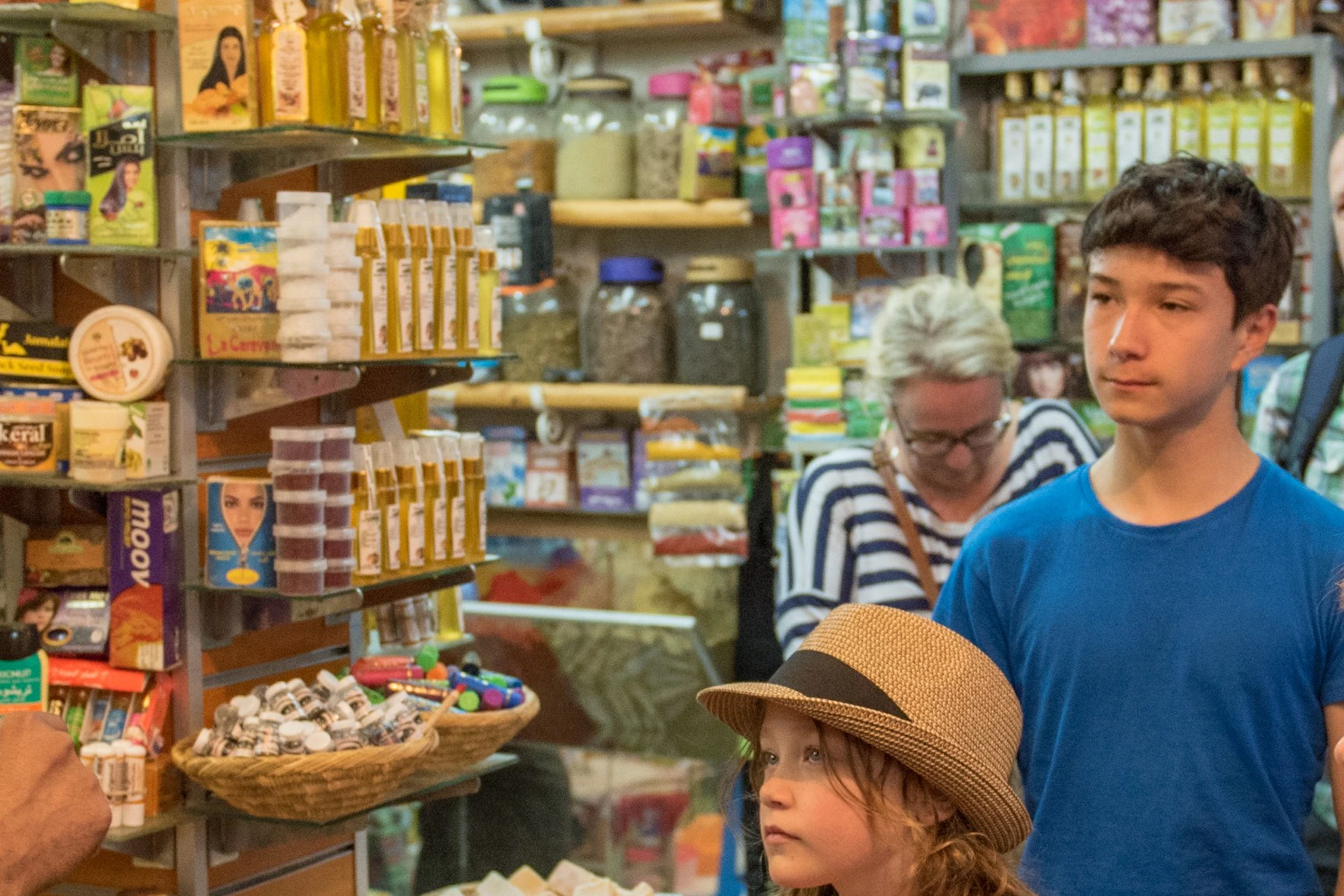 A group of people, including children, stand attentively in a colorful, crowded market shop as a vendor talks to them.