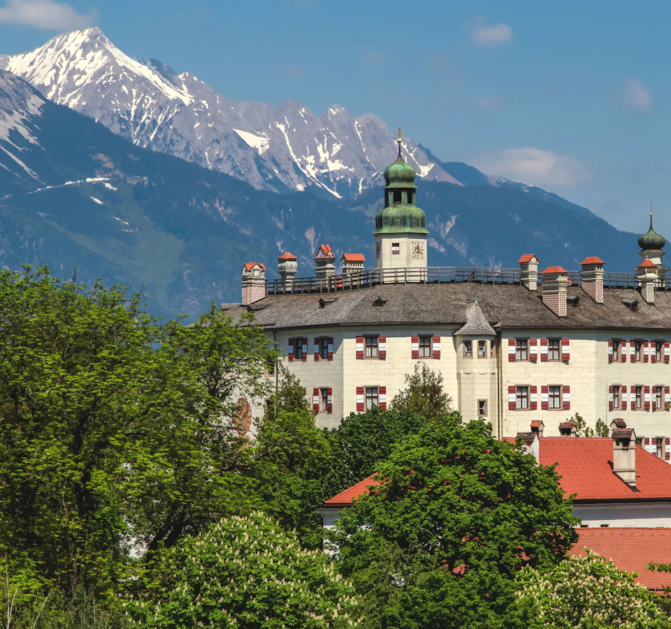 A historic building with green dome towers and red roofs, surrounded by lush trees, with snow-capped mountains in the background.