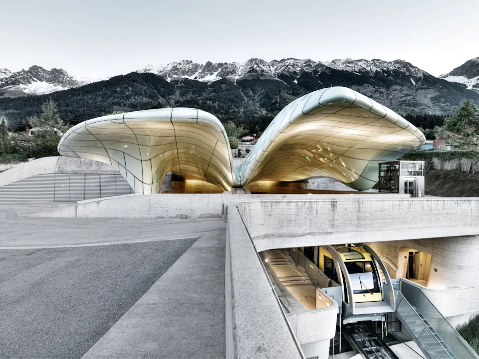 Futuristic, wavy-roofed building with glass and metal structures; mountains in the background, and modern interior spaces visible.