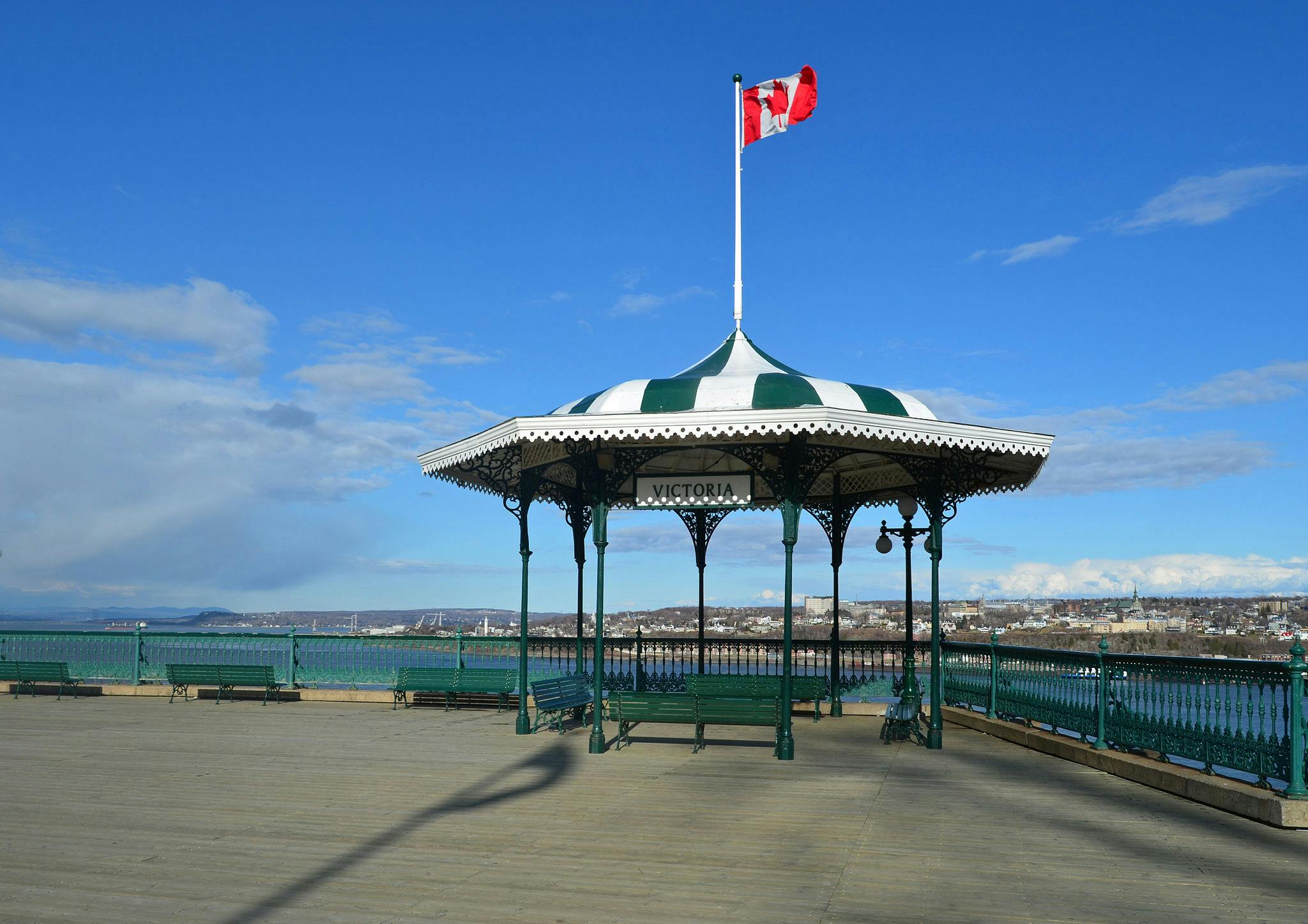 Ένα κιόσκι με το όνομα «Βικτώρια» με καναδική σημαία, πράσινους φράχτες και παγκάκια σε μια ξύλινη πλατφόρμα με θέα σε μια λίμνη και ένα μακρινό αστικό τοπίο.