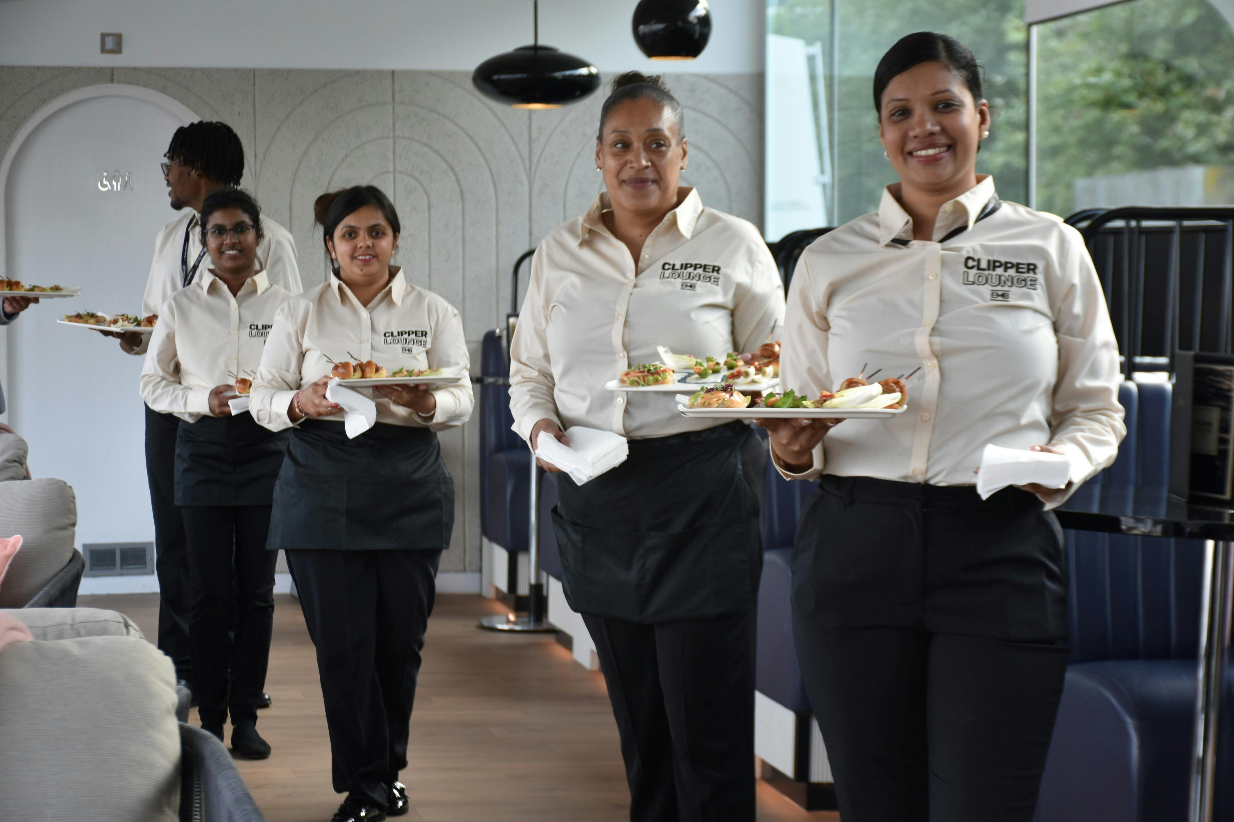 制服を着た3人の笑顔のサーバーが、青いブースと大きな窓があるレストランに食べ物の皿を置いています。