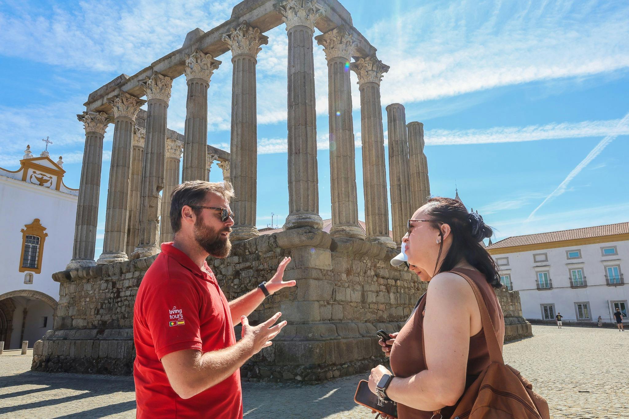 A man in a red polo shirt explains a site to a woman near ancient stone columns under a clear blue sky.